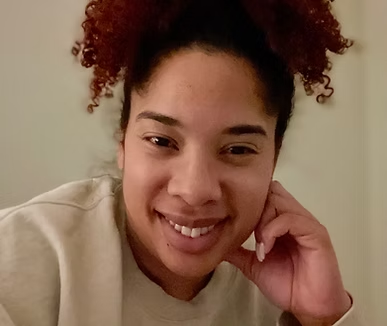 A person with curly hair pulled up, wearing a light-colored sweatshirt, smiles while resting their chin on their hand.