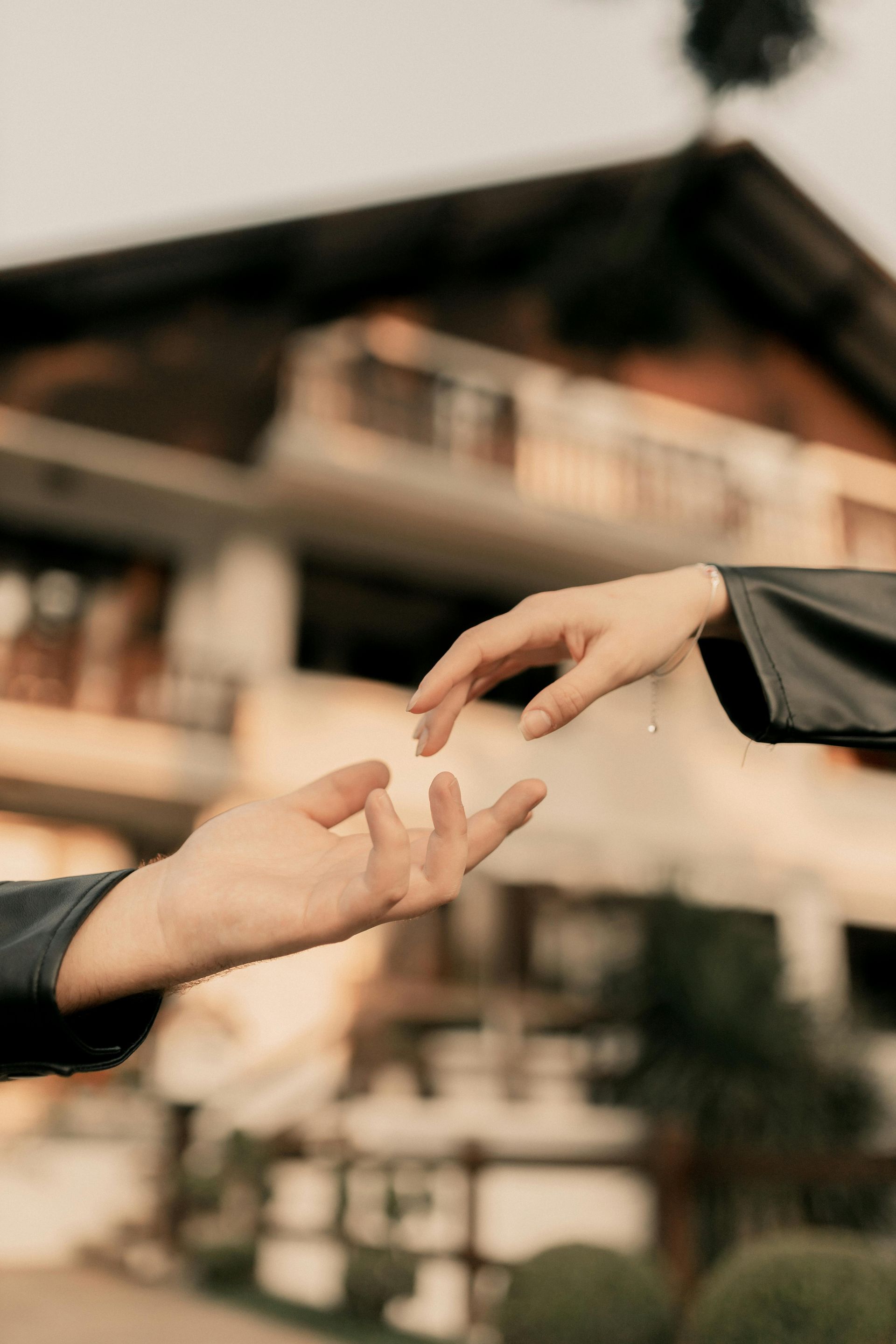 Two hands reaching toward each other in a soft, sunlit setting with a blurred building in the background.