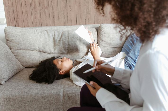 A therapist holding a tablet writes notes while a person lies on a couch, holding a piece of paper.