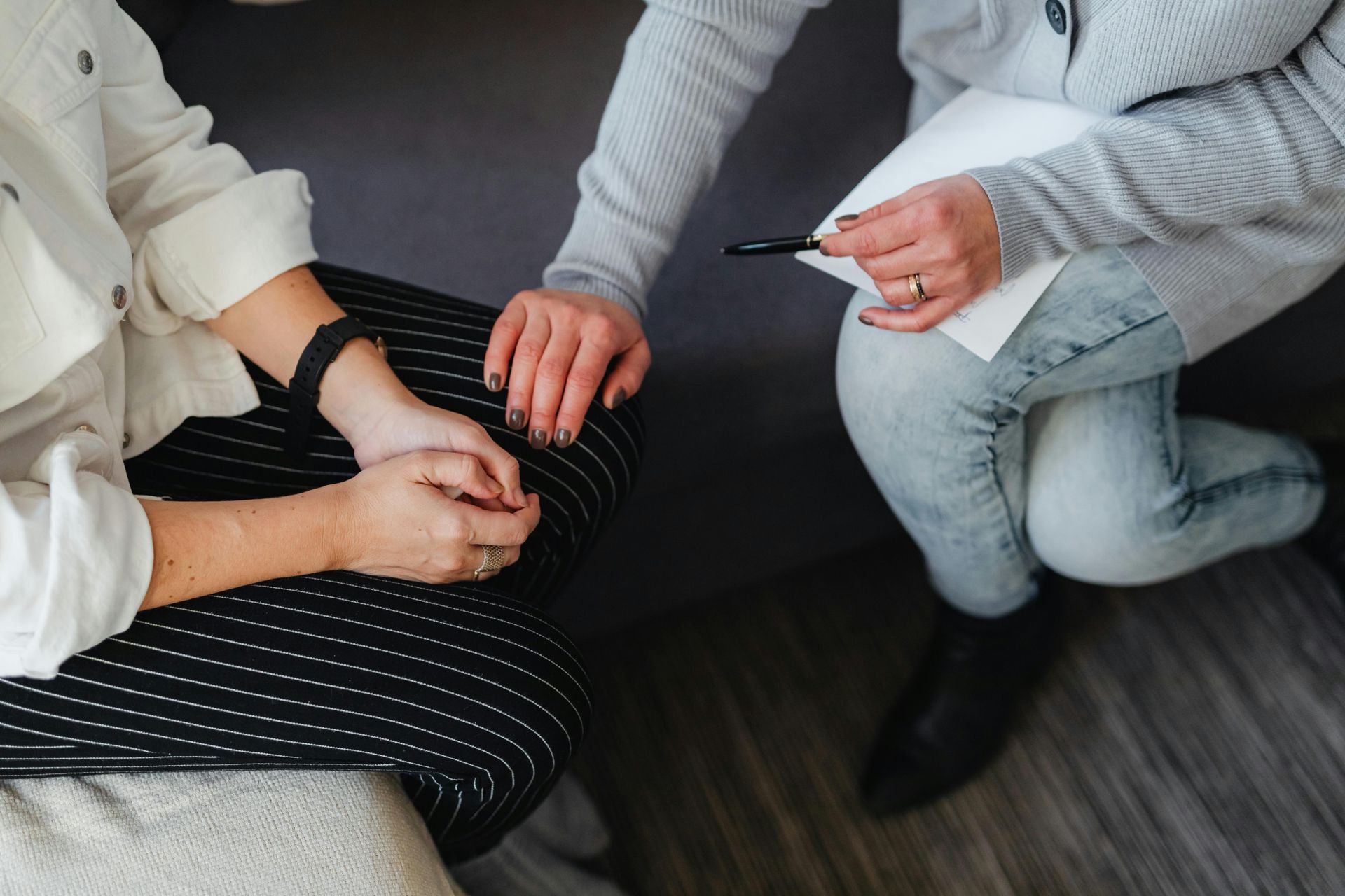 Two people sit closely together, one placing a hand on the other's lap while holding a pen and paper.