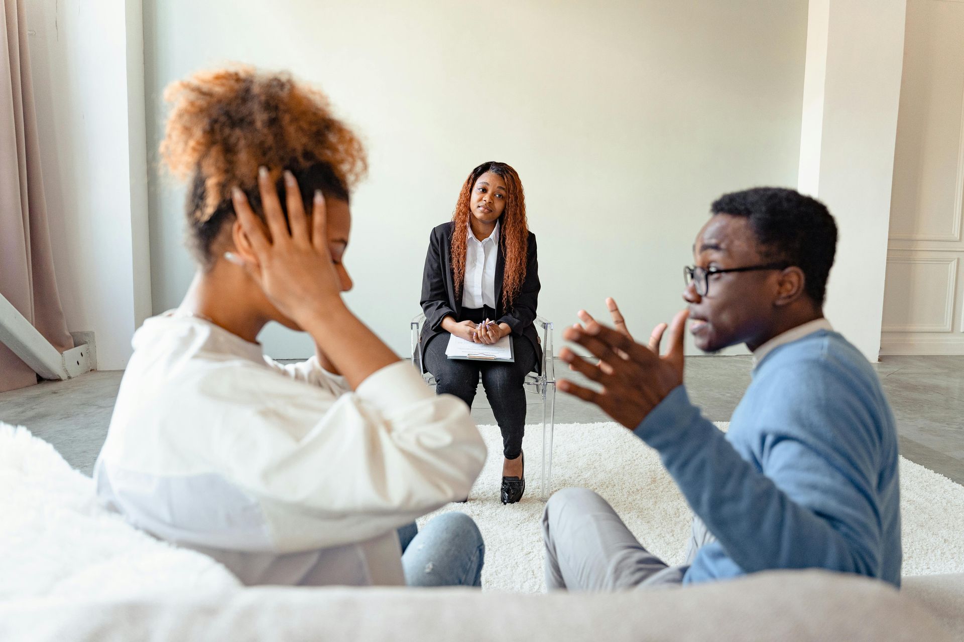 A couple has a tense discussion while seated on the floor in front of a therapist in an office setting.