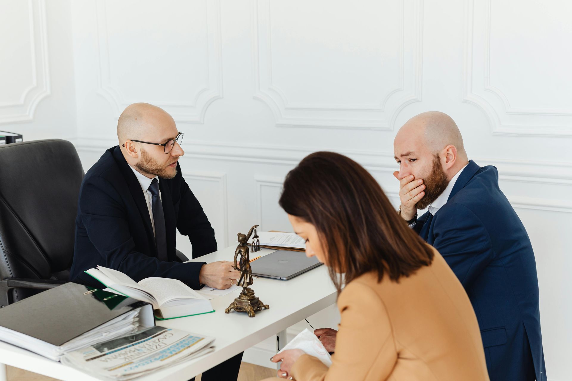 A lawyer sits across a desk from two people in a professional office, featuring a statue of Lady Justice.