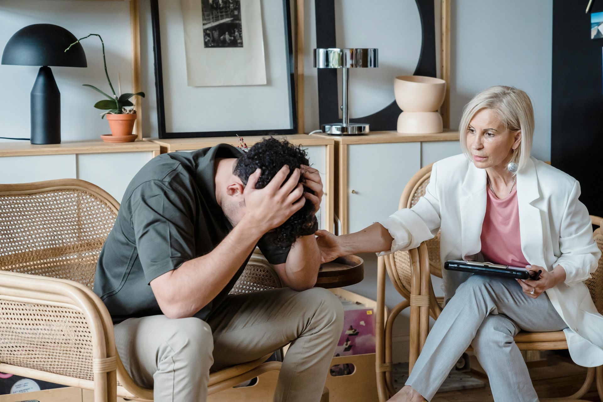 Upset person sitting with head in hands while a counselor reaches out in a bright living room.
