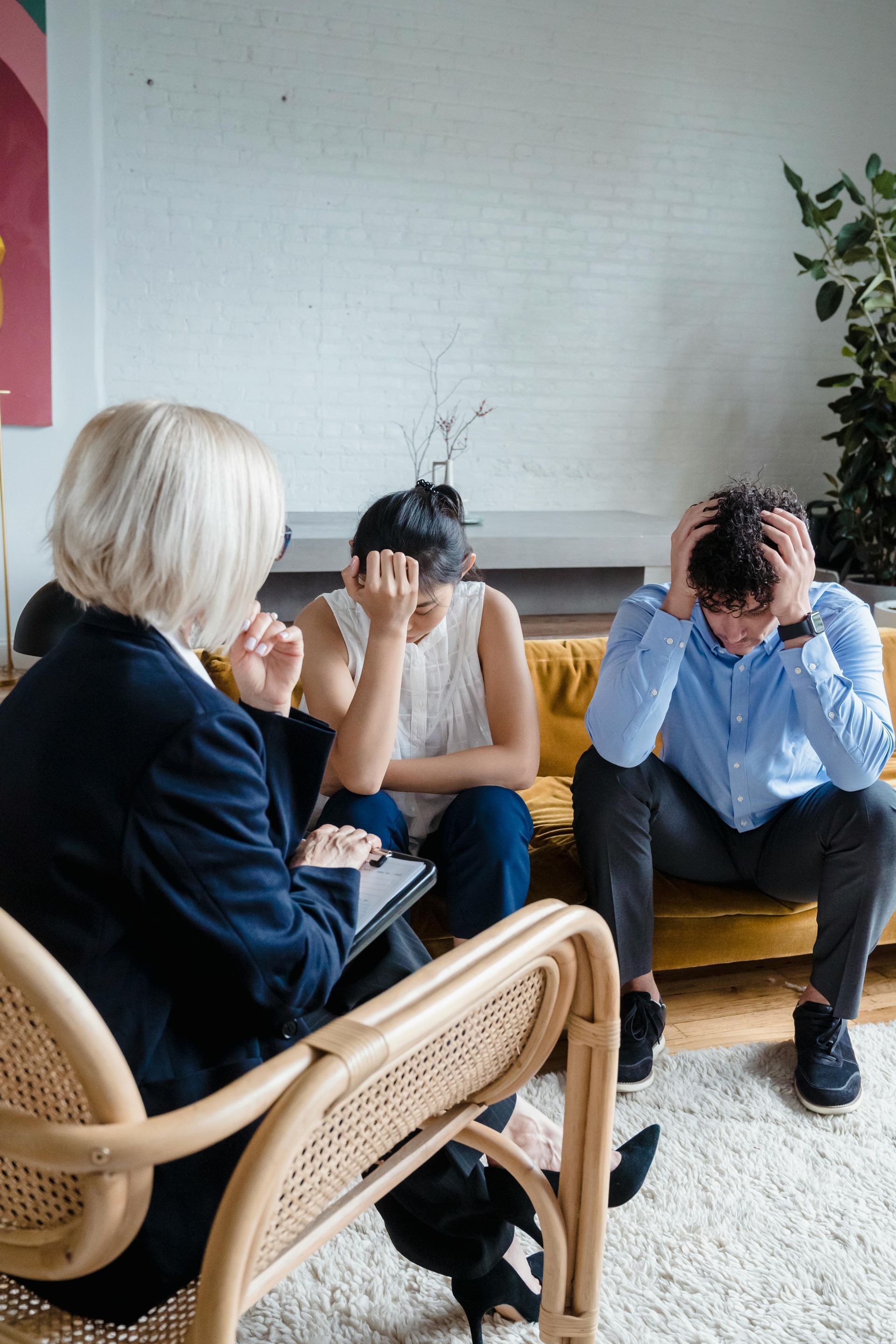 A therapist sits in a chair facing two distressed people on a couch who have their heads in their hands.