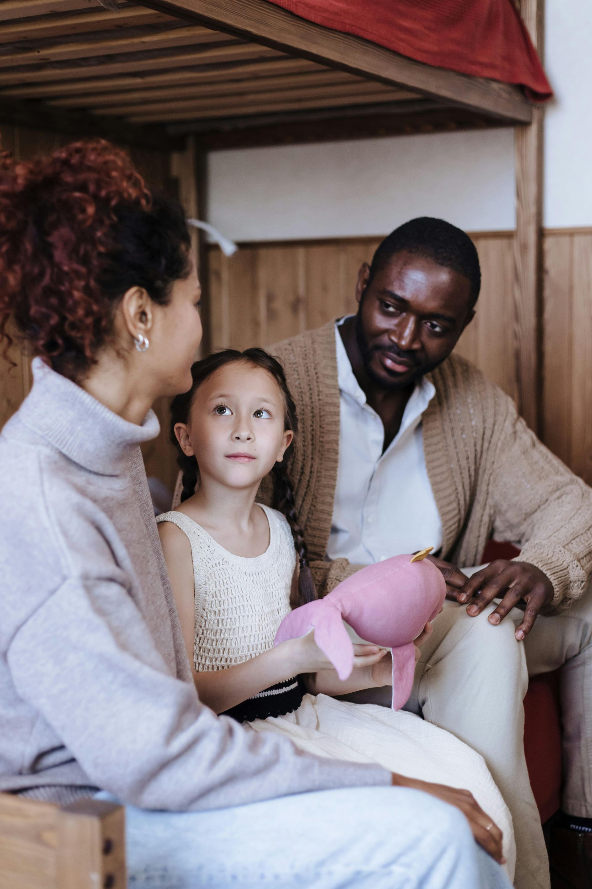 A parent and child sit together indoors, with the child holding a small pink plush toy while looking toward the parent.