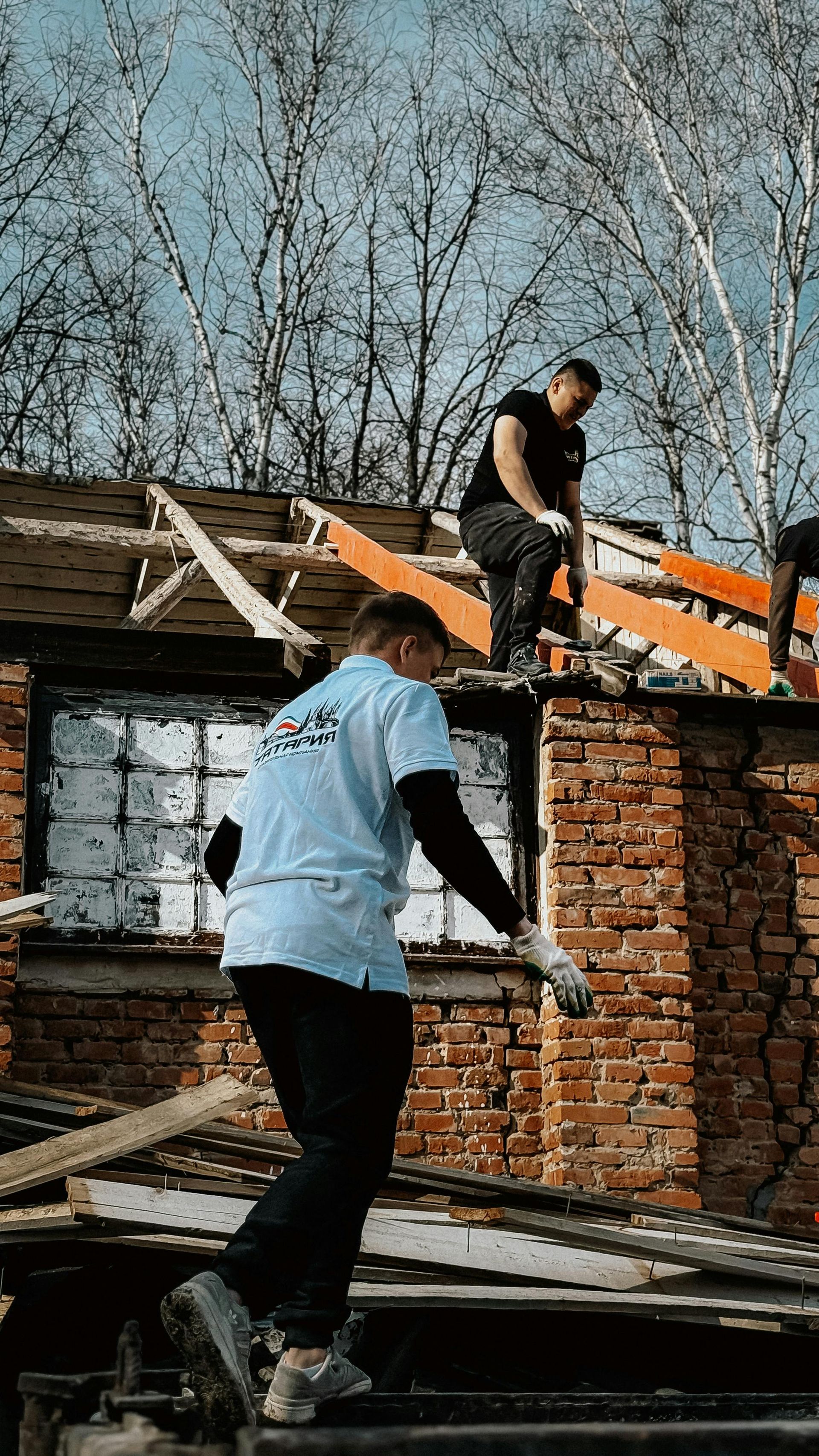 Two people on a damaged brick rooftop, one in a light blue shirt climbing amid scattered debris and broken beams.