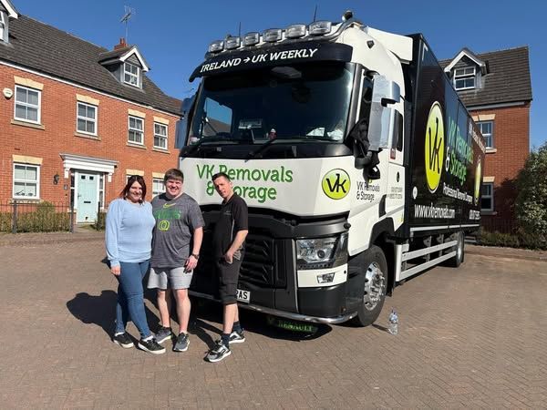 Three people pose with a VK Removals truck in front of houses on a sunny day.