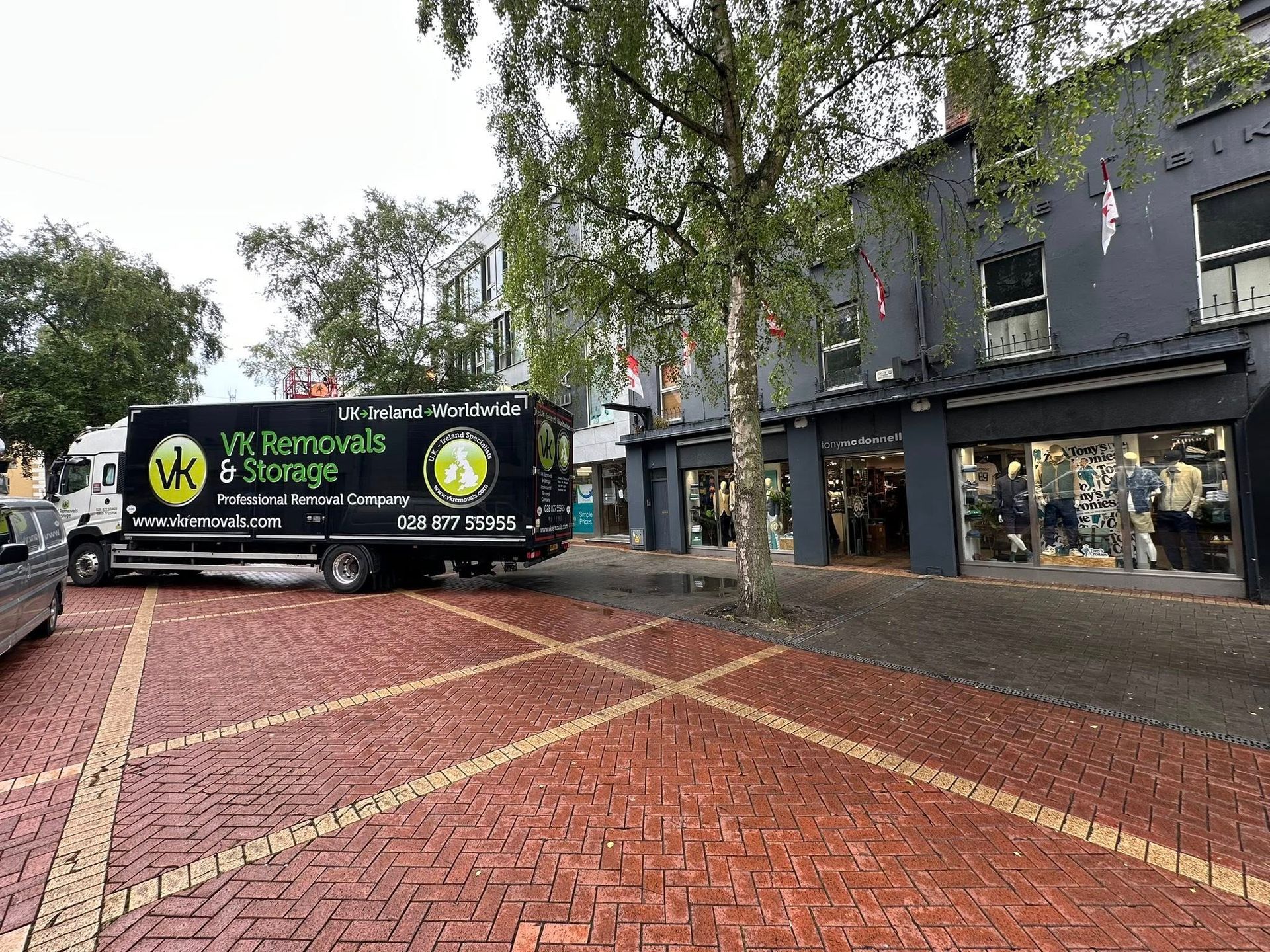 A removal truck parked on brick road next to a store, with trees and buildings in the background.