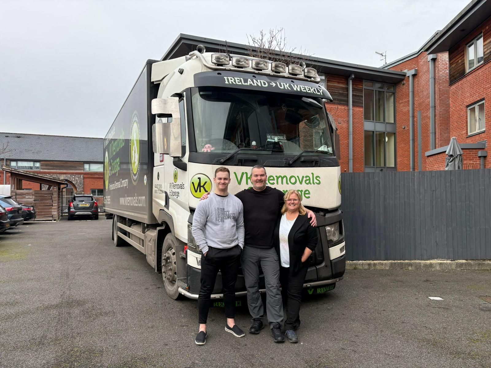 Three people posing in front of a large white moving truck with business branding in a parking lot.