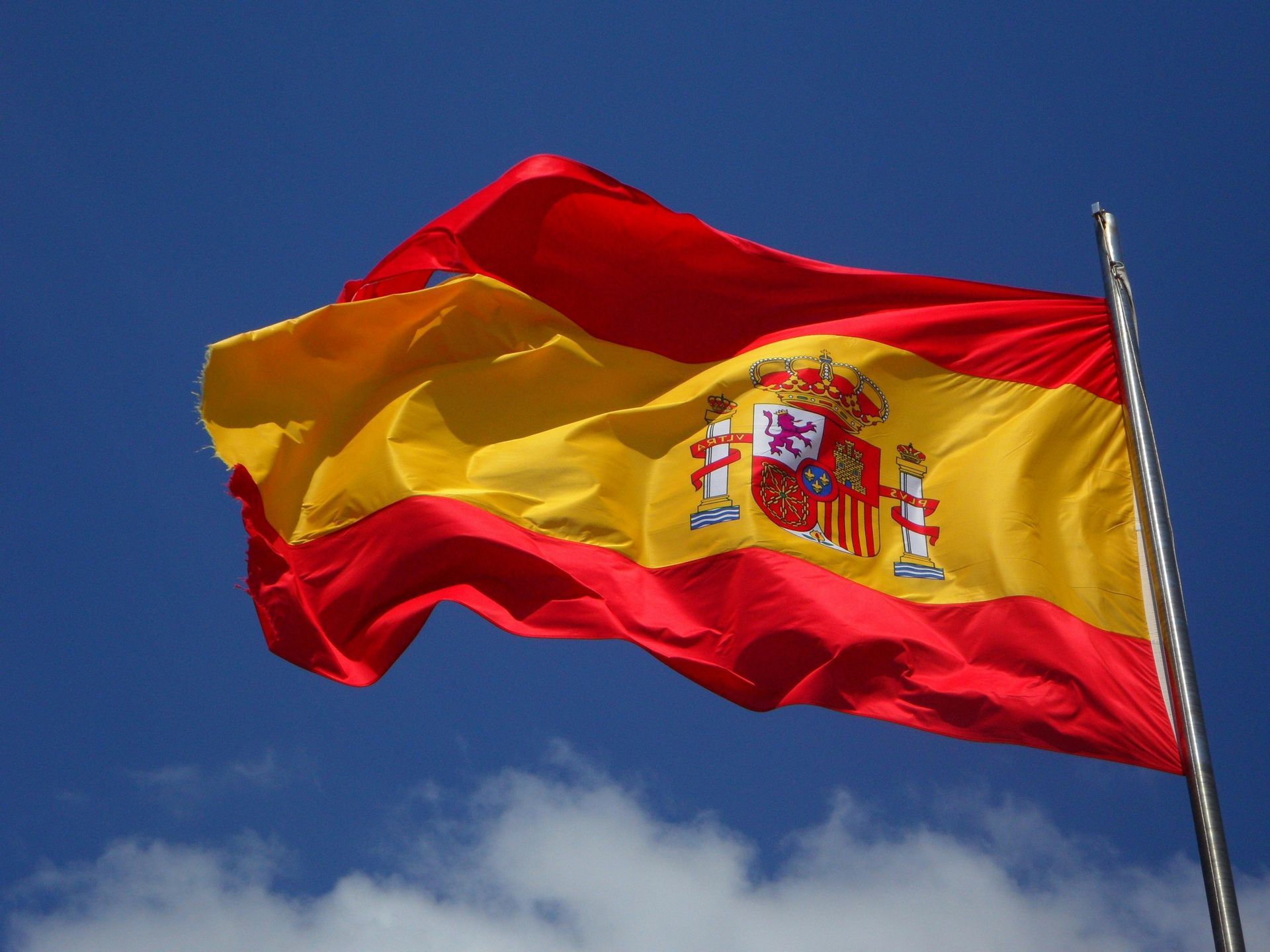Flag of Spain waving in the wind against a blue sky, with the Spanish coat of arms on the yellow stripe.