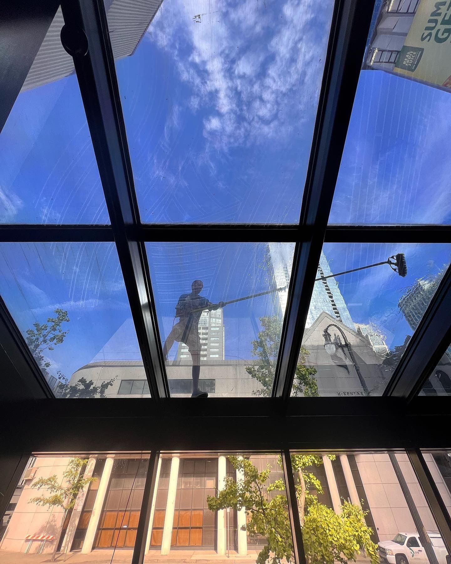 Person cleaning a glass skylight with a long-handled tool, city buildings and blue sky visible.