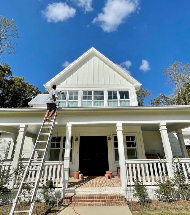 A person on a ladder paints the exterior of a white, gabled house with a front porch under a clear blue sky.