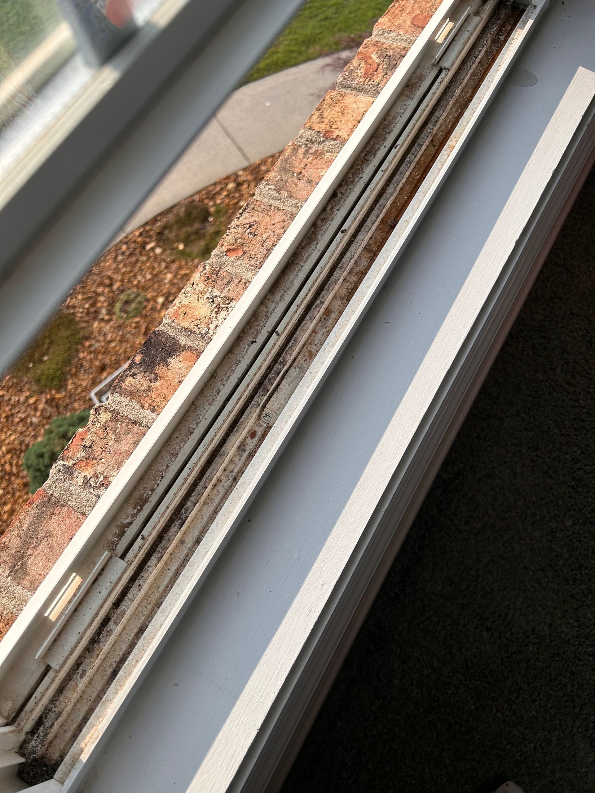 Close-up of a dirty, debris-filled window track on a white vinyl window frame overlooking a brown, mulched garden bed.