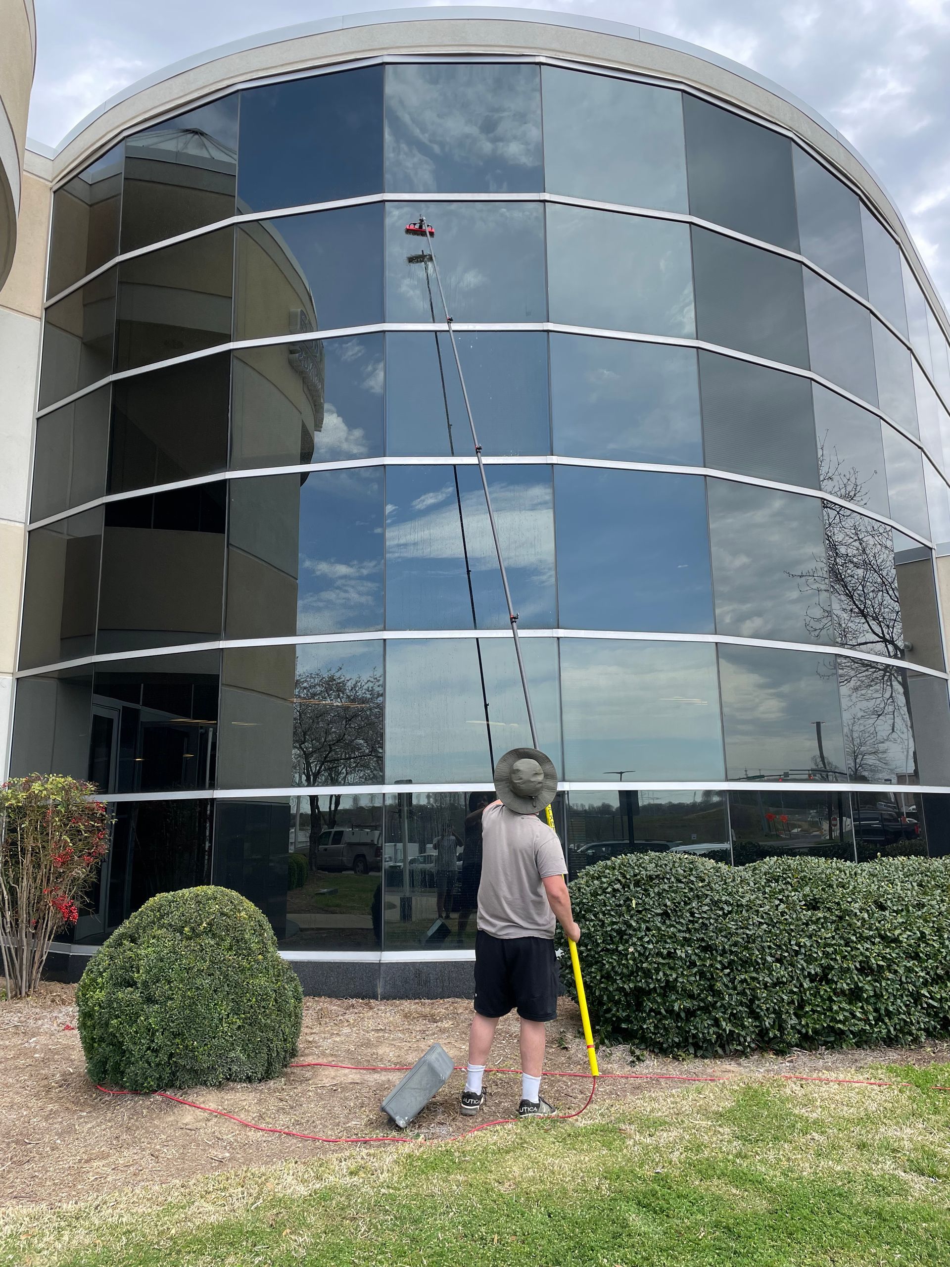 Person washing windows on curved glass building using a long-handled tool.