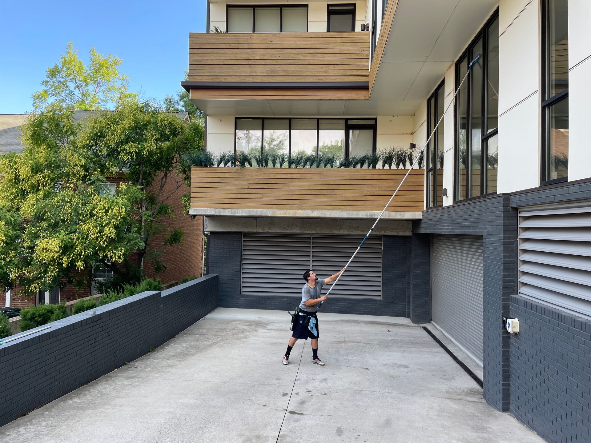 Man using a long pole to clean windows on a multi-story building. Concrete driveway in front.