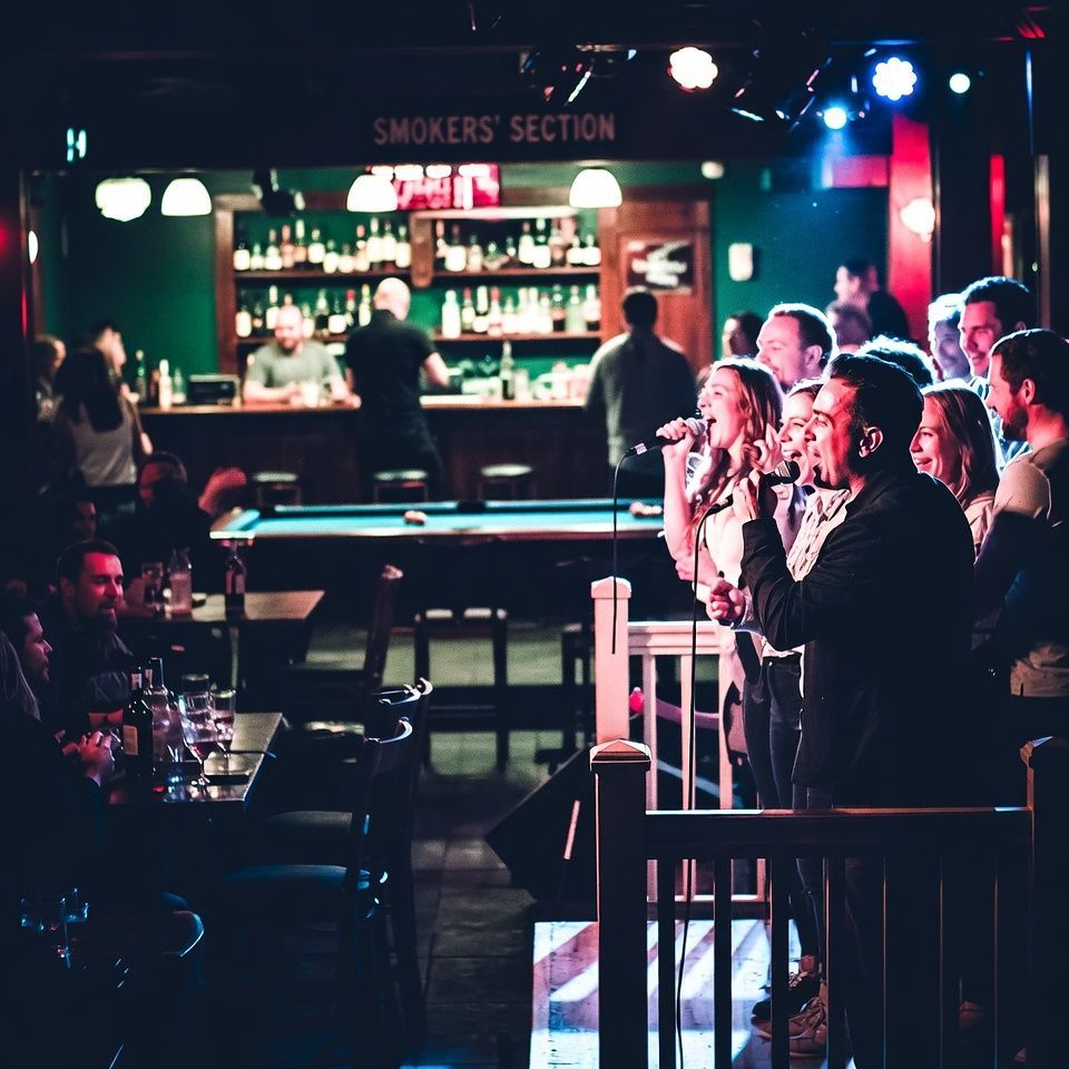 People singing karaoke under a red neon “Karaoke” sign in a lively bar.