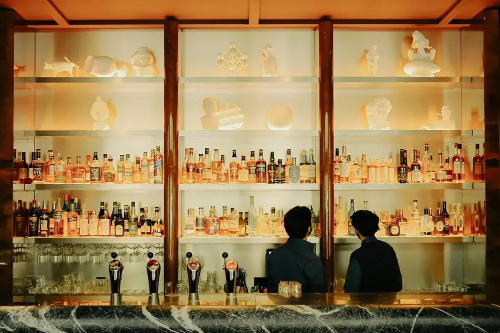 Backlit bar shelves lined with bottles, with two people seated at the counter in front