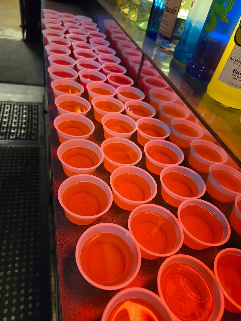 Rows of red plastic cups filled with orange liquid on a bar counter, with bottles lined up behind them