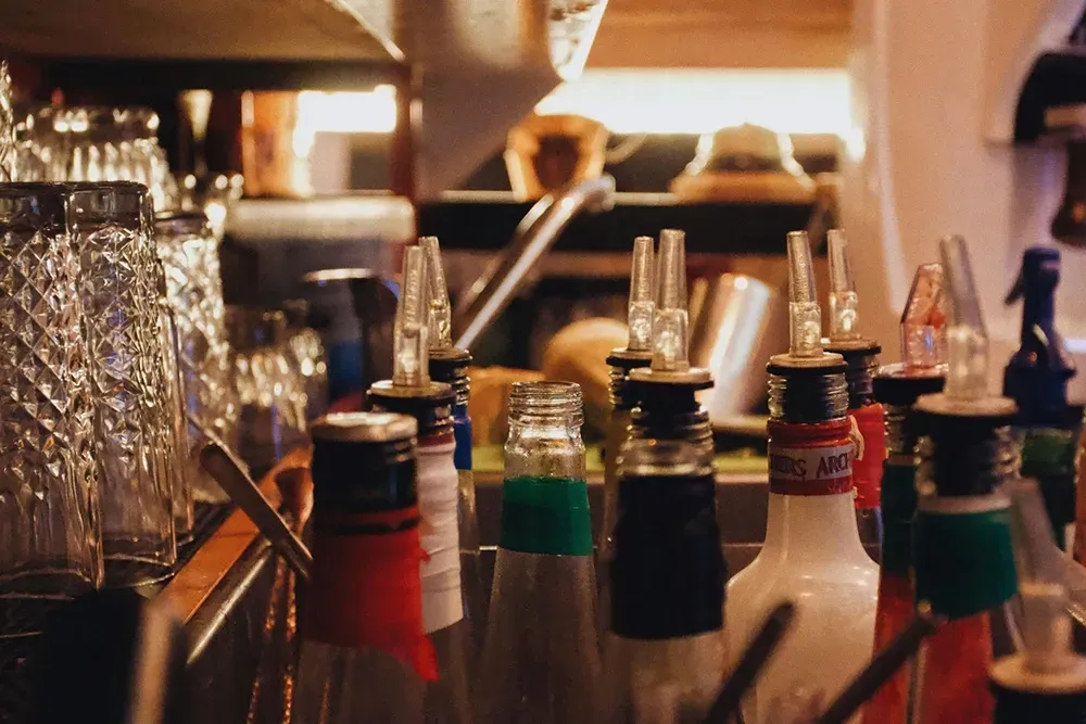 Bar counter lined with assorted cocktail bitters bottles and bar tools in warm light
