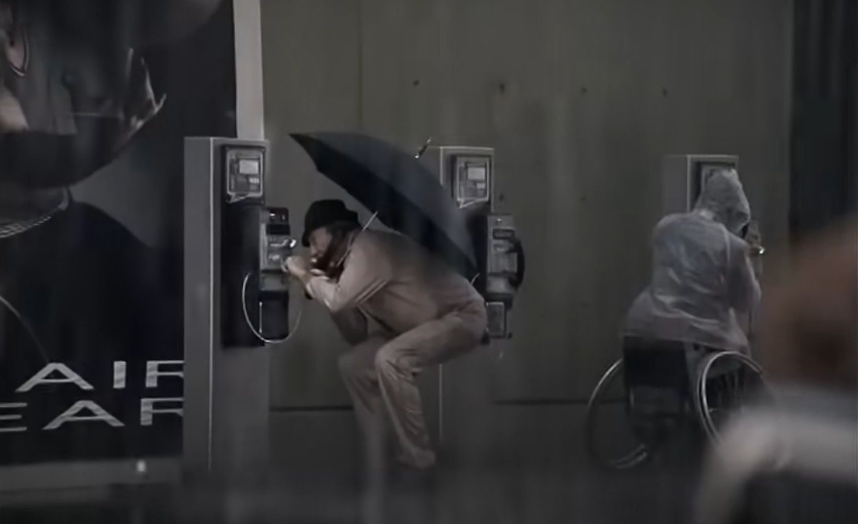 The scene is from a 2000s Electricite de France advertisement. A moody, rainy street with two people using public payphones. One man in a trench coat and hat crouches uncomfortably under a large black umbrella while speaking on the phone. To his right, a person in a wheelchair, covered in a transparent rain poncho, is also using a payphone. The wall behind them is bare concrete, and a large poster partially visible on the left adds to the urban atmosphere. It highlights the experience of people with disabilities as a product of the choices that were made when building the environment that they live in.
