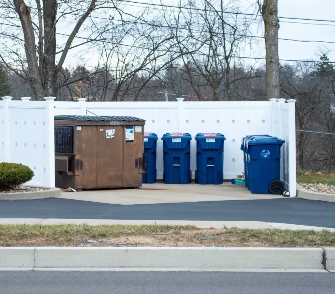 A large brown dumpster and four blue recycling bins enclosed by a white vinyl privacy fence outdoors.