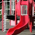 A red plastic playground slide attached to a metal structure with a white spiral pole on a paved play area.
