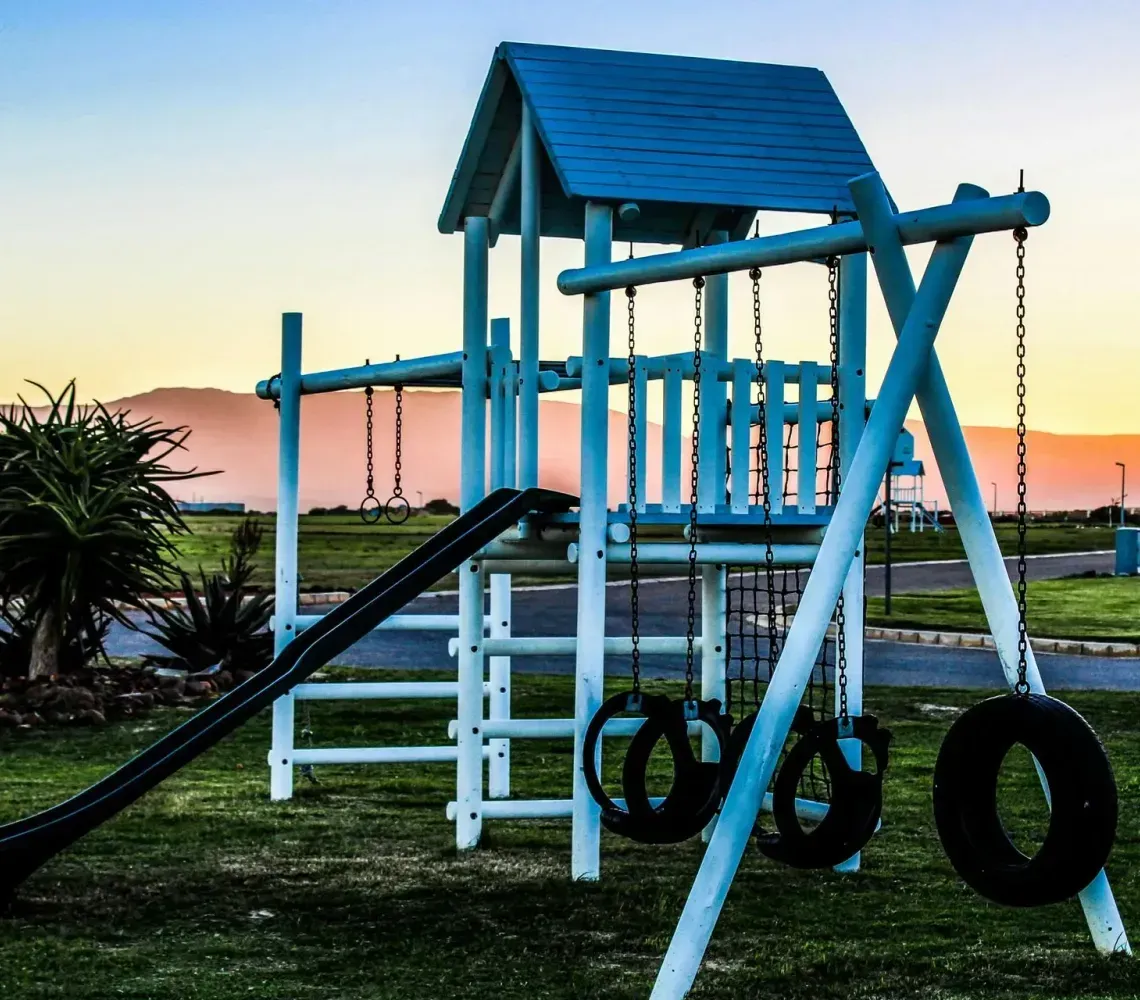 A blue wooden playground structure with a slide and tire swings at sunset in a grassy park.