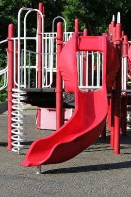 A red plastic playground slide attached to a metal structure on a paved surface.