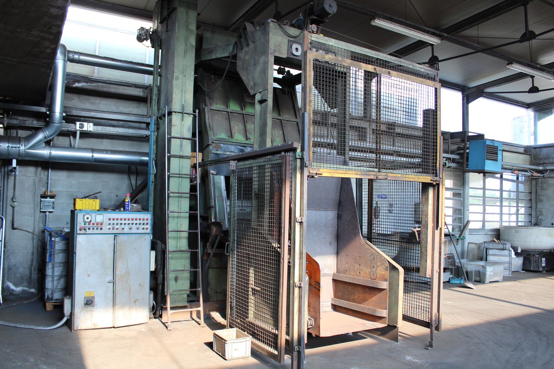 Macchinario industriale in una fabbrica, con gabbia metallica, nastro trasportatore e quadro di controllo sotto tubi aerei.