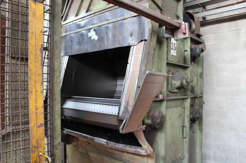 Macchinario industriale in metallo danneggiato con scivolo aperto accanto a un muro di cemento e a una recinzione di sicurezza.