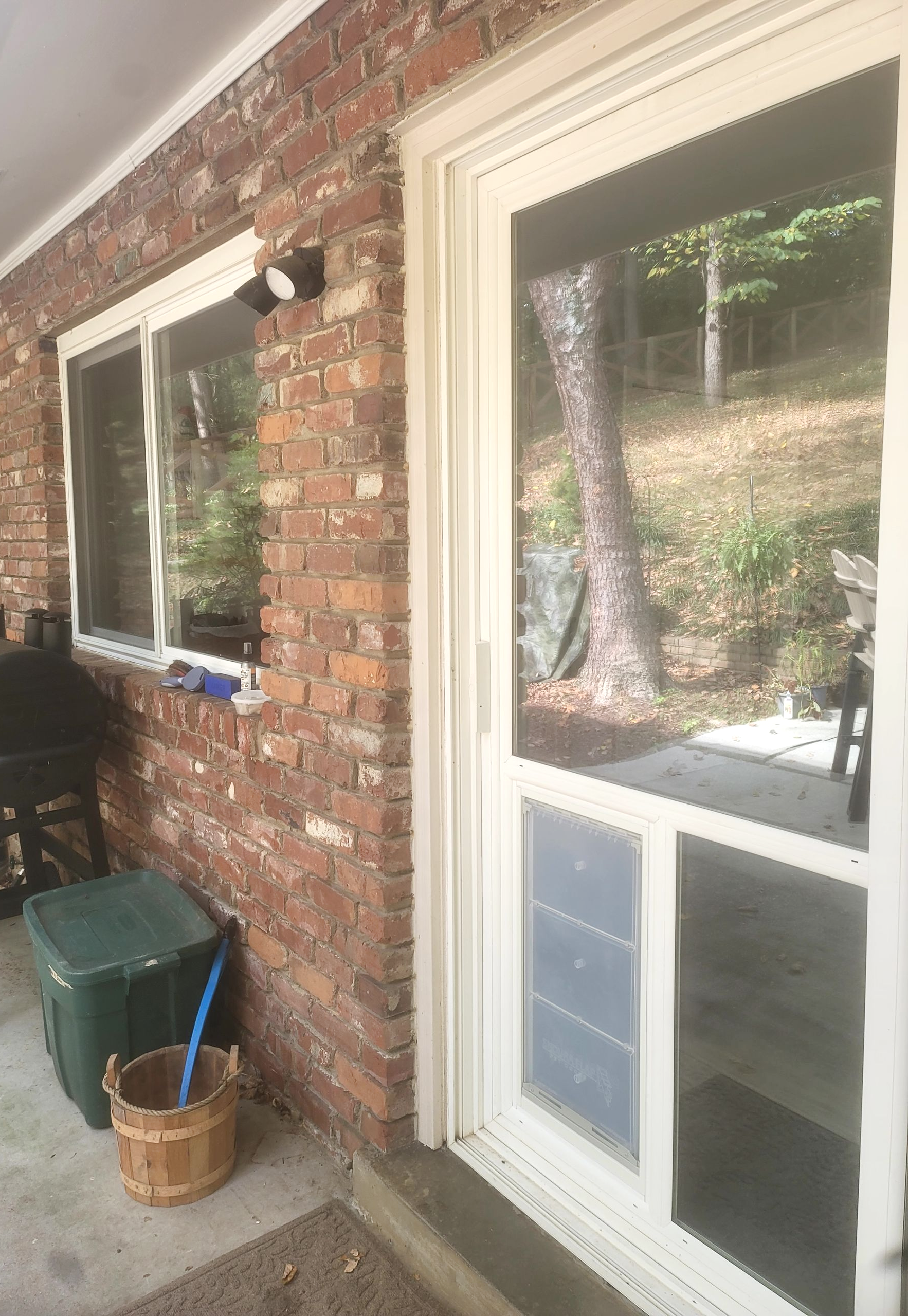 A white screen door with a pet door, set in a rustic brick exterior wall beside a window, with an outdoor area visible.