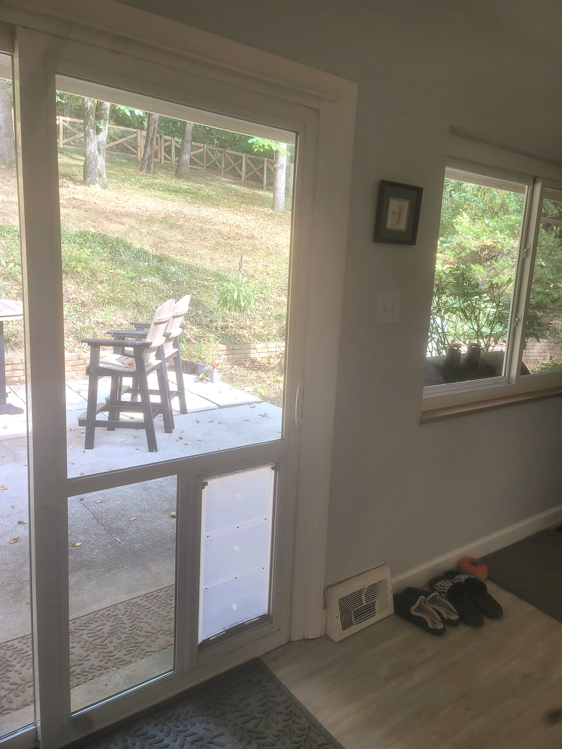 A sliding glass door with a built-in pet door, looking out onto a patio with chairs and a wooded area.