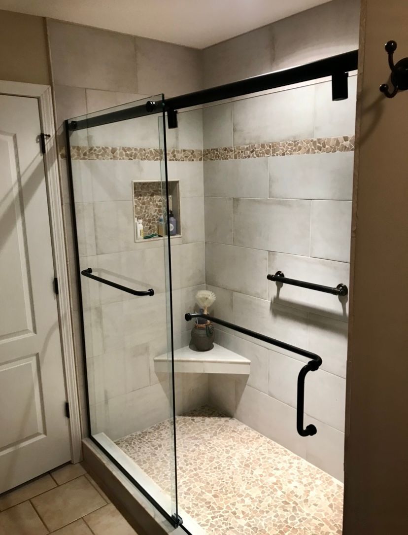 A walk-in tiled shower featuring a corner bench, a recessed shelf, and matte black grab bars and door hardware.