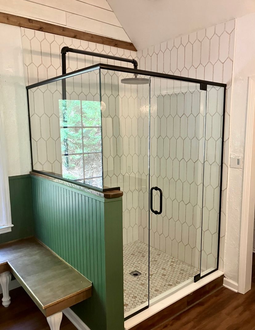 A walk-in shower with a black frame and white elongated hexagonal tile, set next to a green paneled wall and bench.
