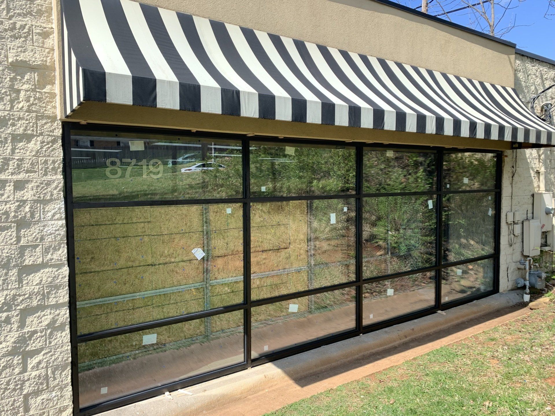 A storefront exterior with large black-framed windows, a brick wall, and a black and white striped awning overhead.
