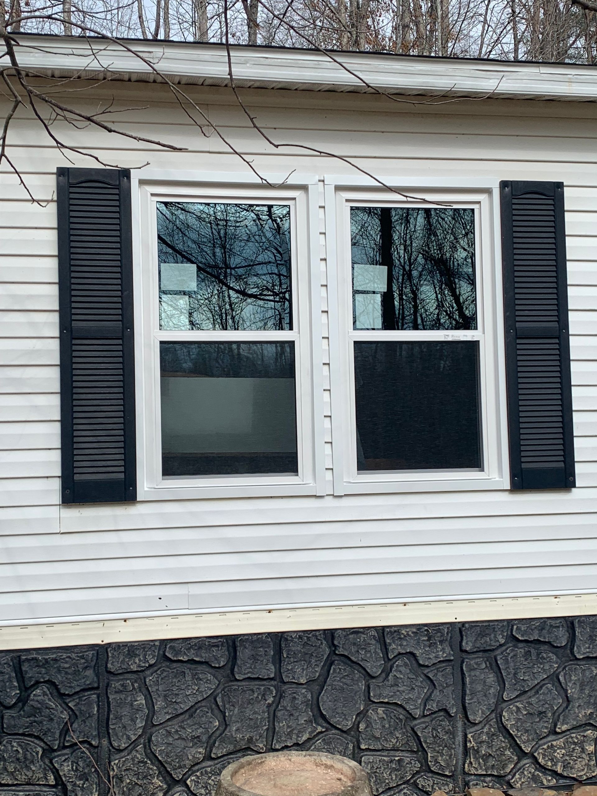 Exterior wall of a home with white horizontal siding, two side-by-side windows with black shutters, and stone foundation.