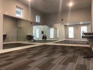 A large, empty fitness room with wall-to-wall mirrors, light grey walls, and brown patterned carpet flooring.