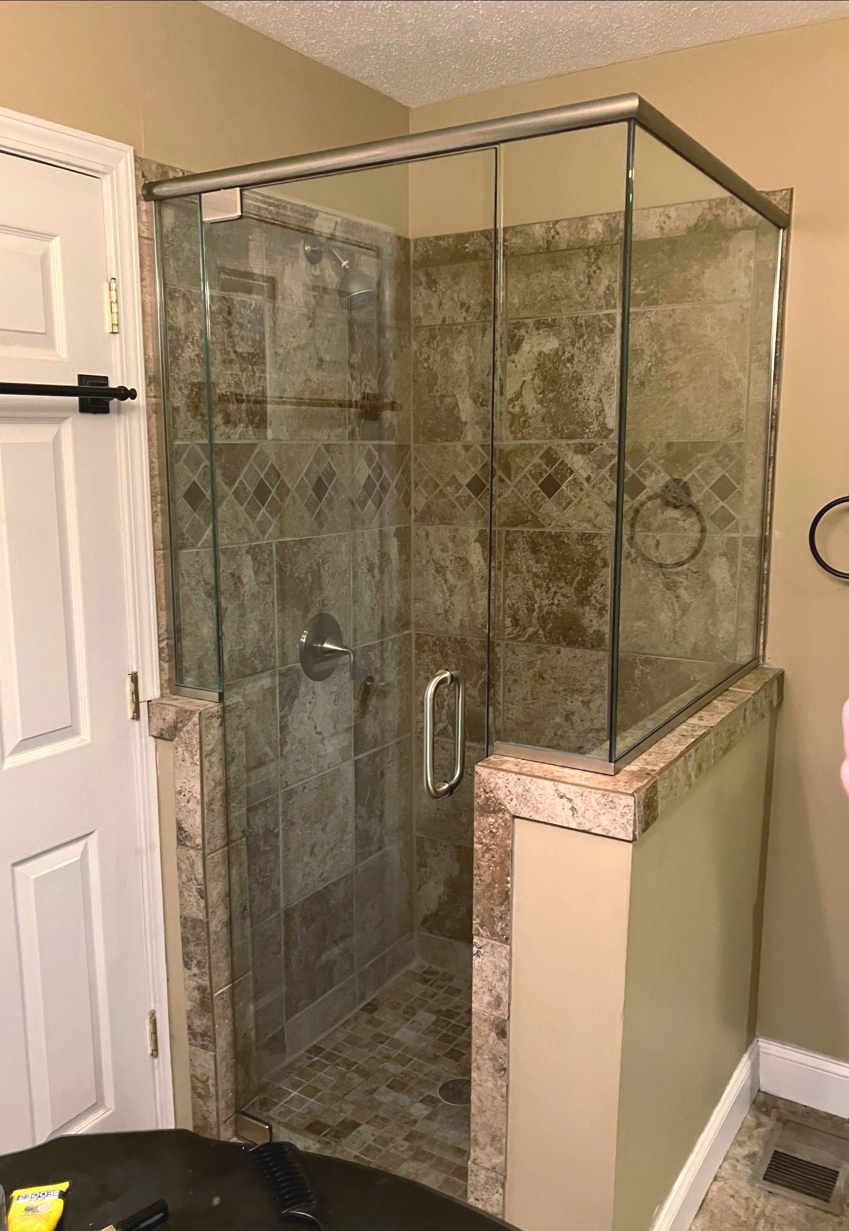 A walk-in shower with a glass door, framed in silver, set against tan stone tile walls and a half-wall ledge.