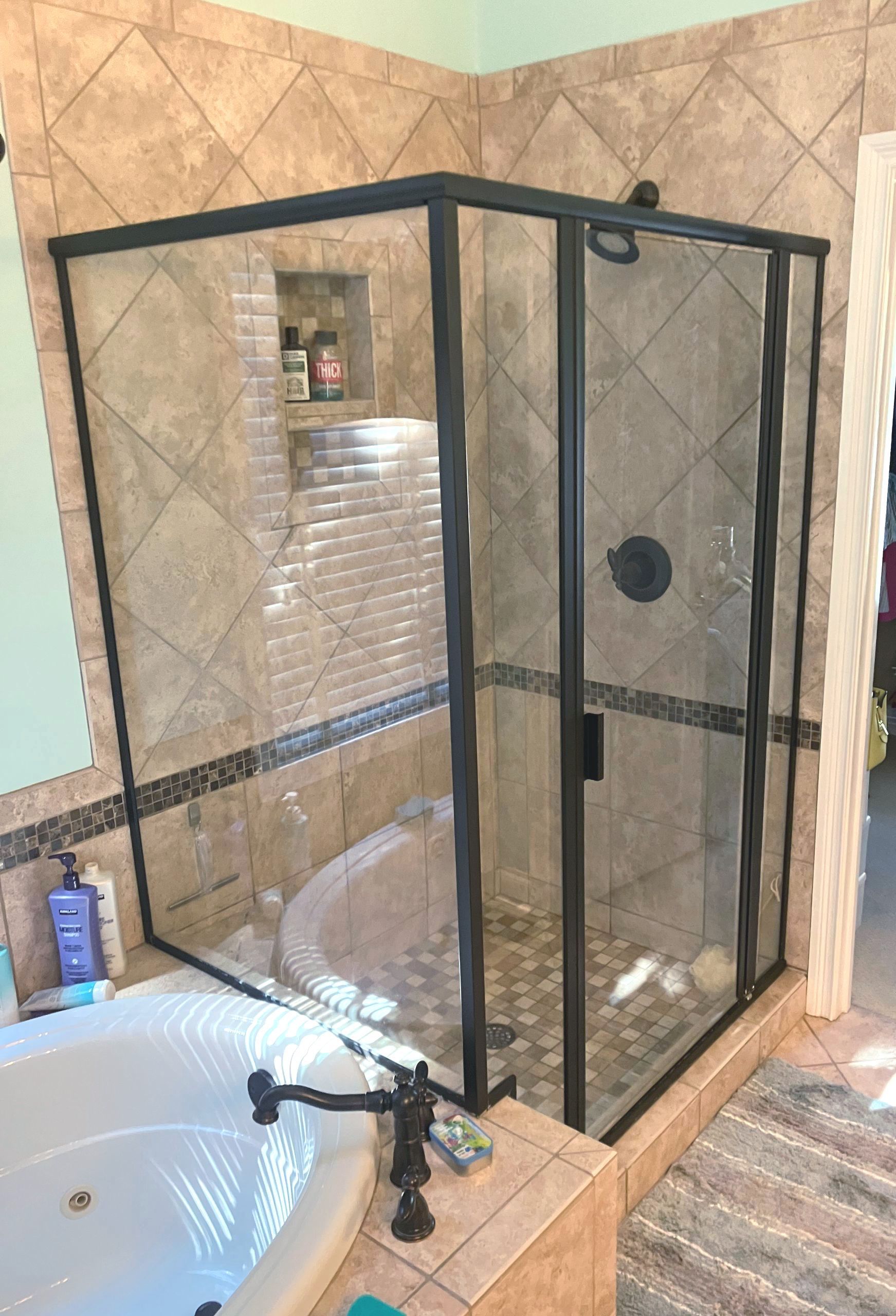 A corner glass shower stall with black frames, surrounded by tan tile walls, situated next to a white bathtub.