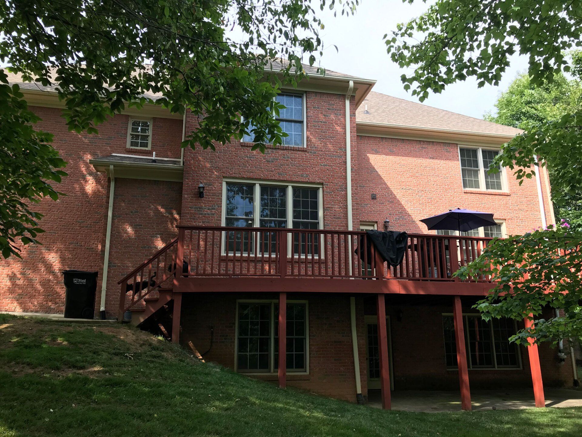 Two-story red brick house with a wooden deck and basement walkout, surrounded by trees on a sloped grassy yard.