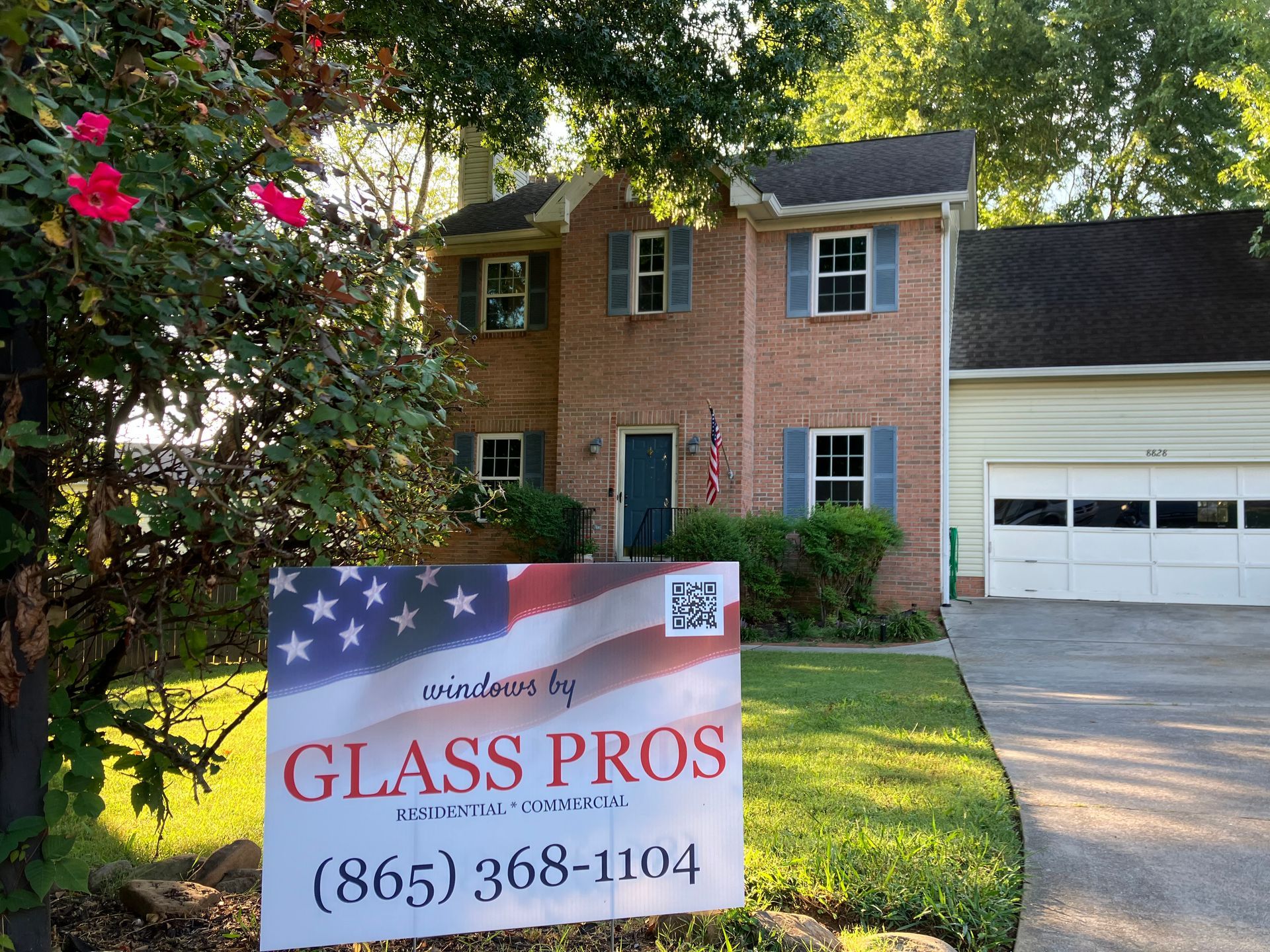 A yard sign for Glass Pros with a US flag design stands in front of a two-story brick house with a garage.