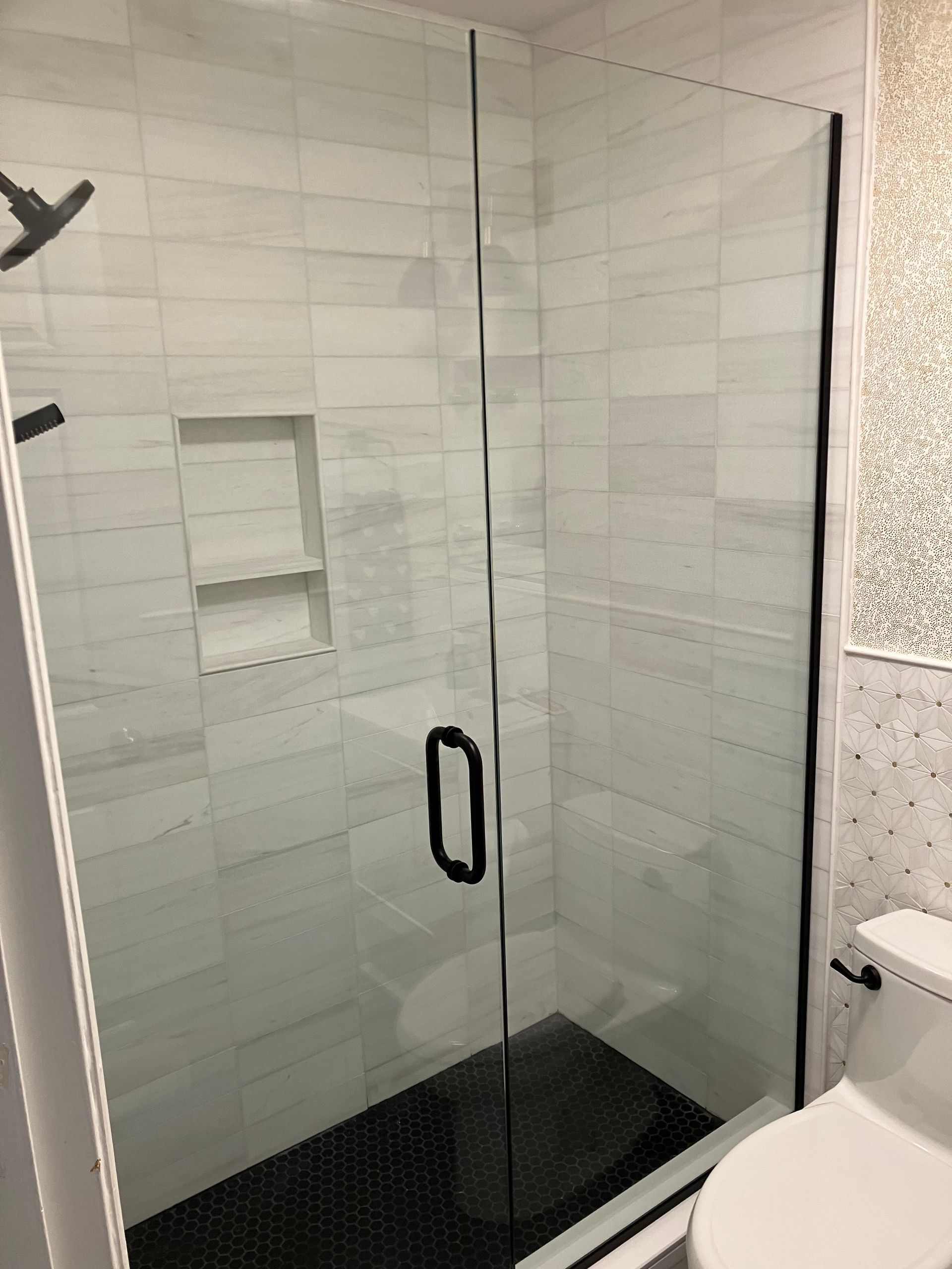 A modern glass-enclosed shower with white tiled walls, a built-in niche, black hardware, and black pebble floor tiling.