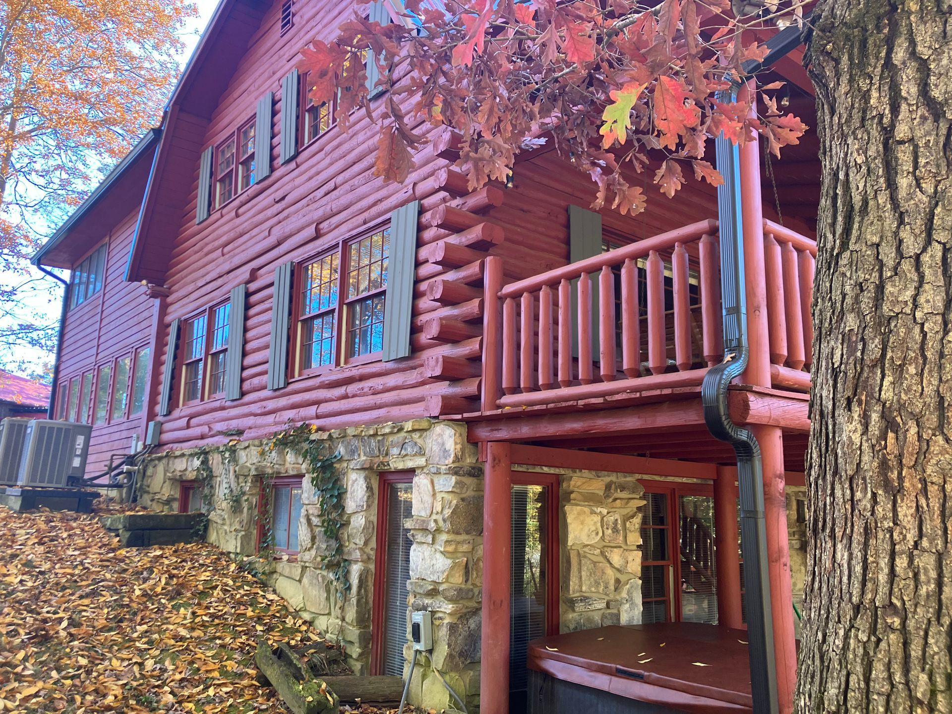 A rustic, red log cabin with a stone foundation, set among fallen autumn leaves with a porch under a large tree.