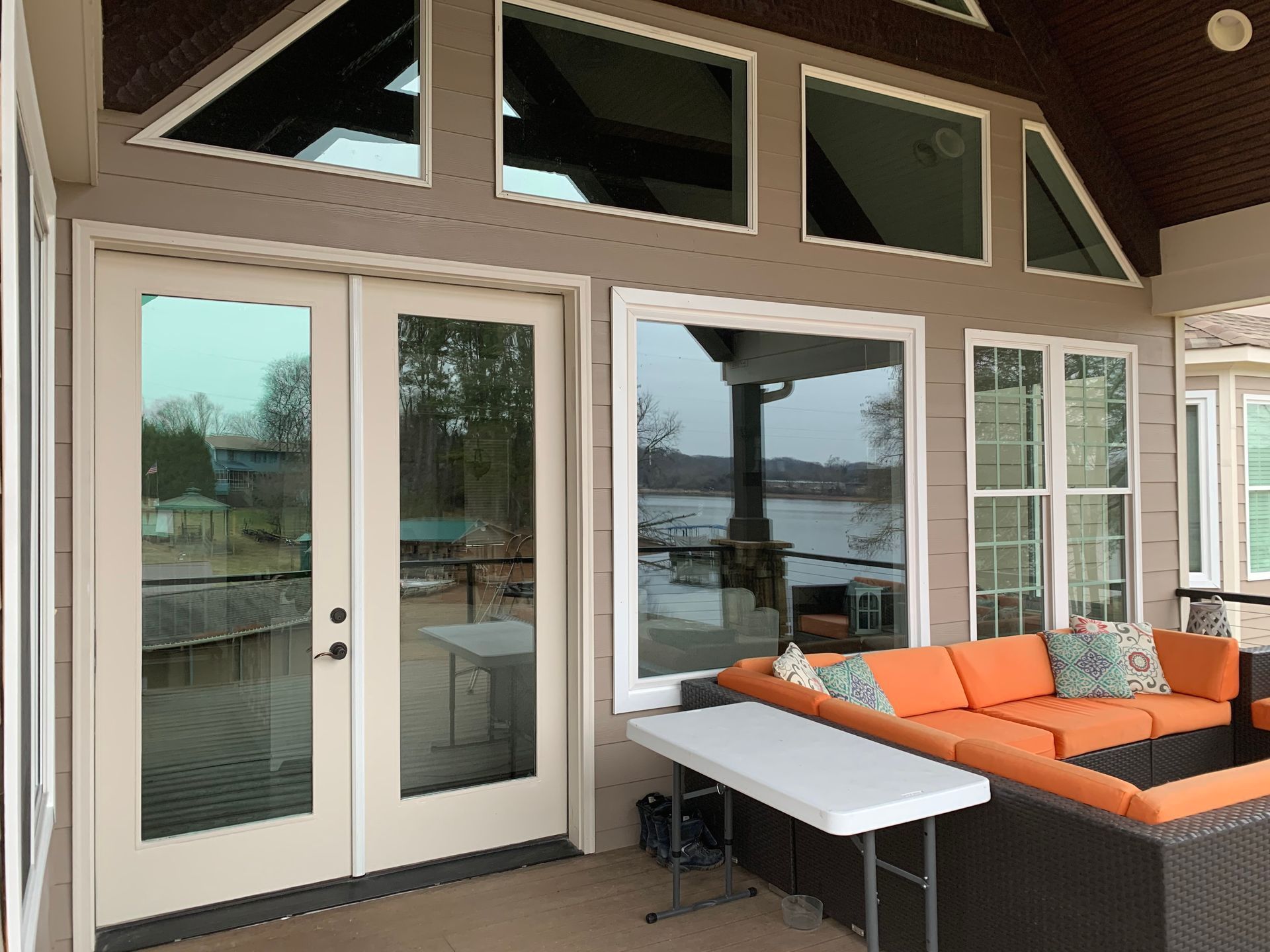 A porch features a glass double door, large windows, and a sectional sofa with orange cushions facing a lake view.