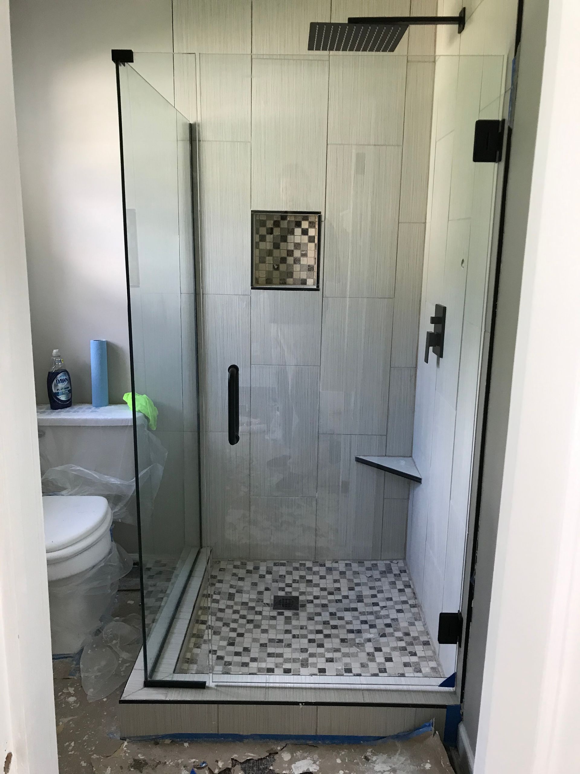 A glass-enclosed shower with light gray tiles, a patterned mosaic floor and inset shelf, and black fixtures.
