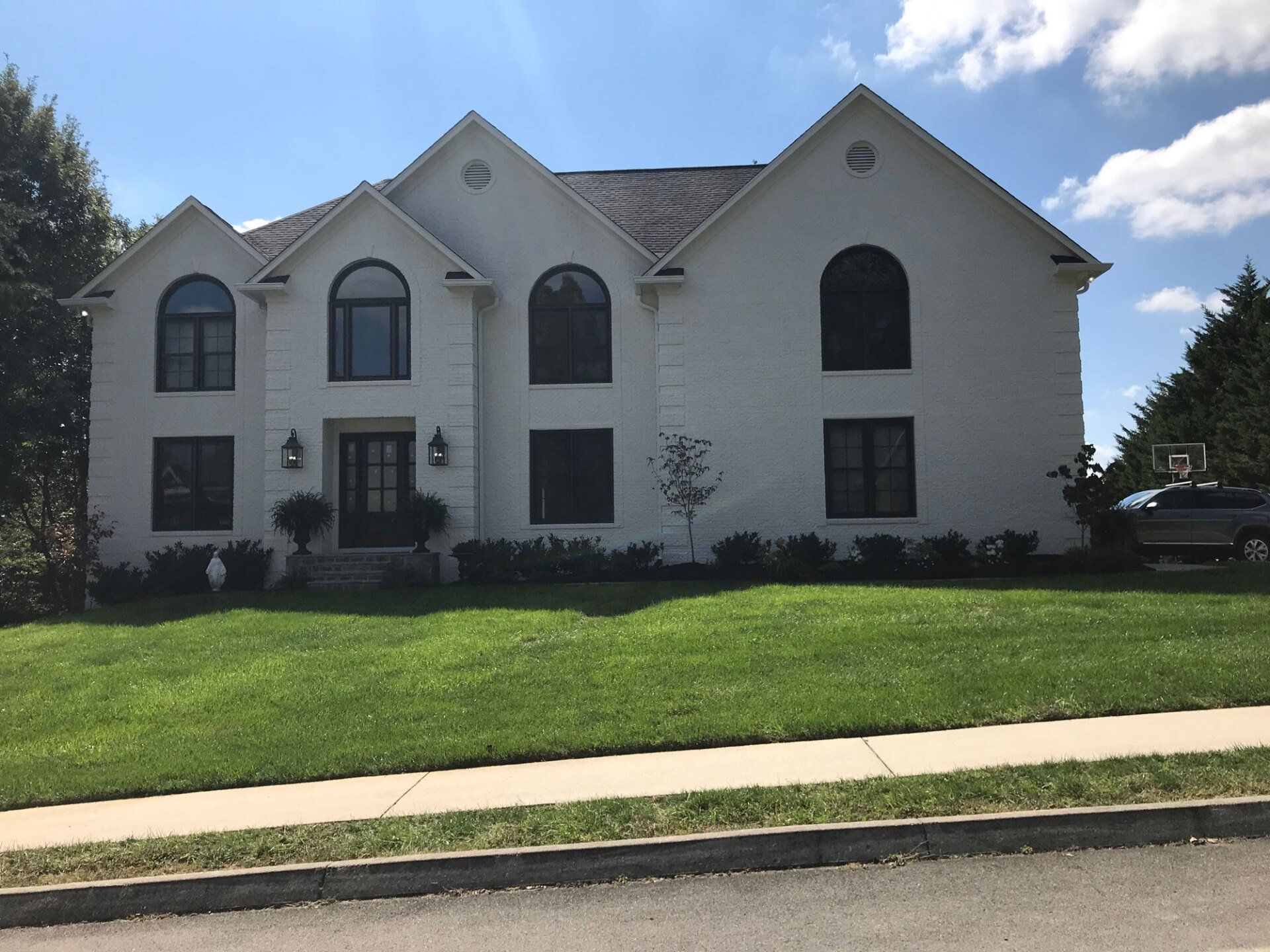 A two-story, white brick house with dark trim, arched windows, and a manicured lawn under a sunny, partly cloudy sky.