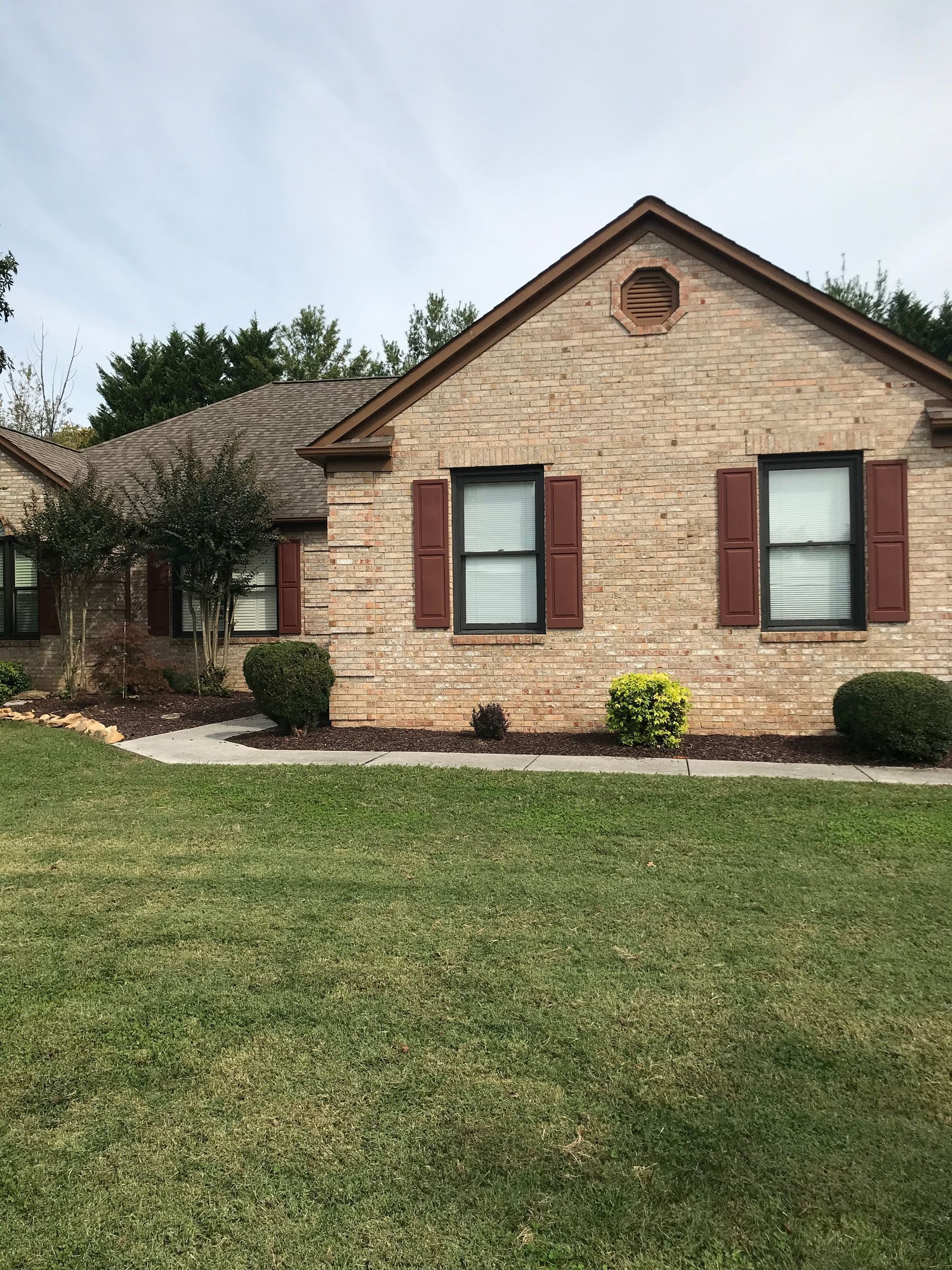 A single-story tan brick house with dark brown shutters, a gabled roof, and landscaped bushes in the front yard.
