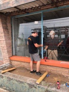 Two people stand outside a building, working on the installation of a large glass window pane within a metal frame.