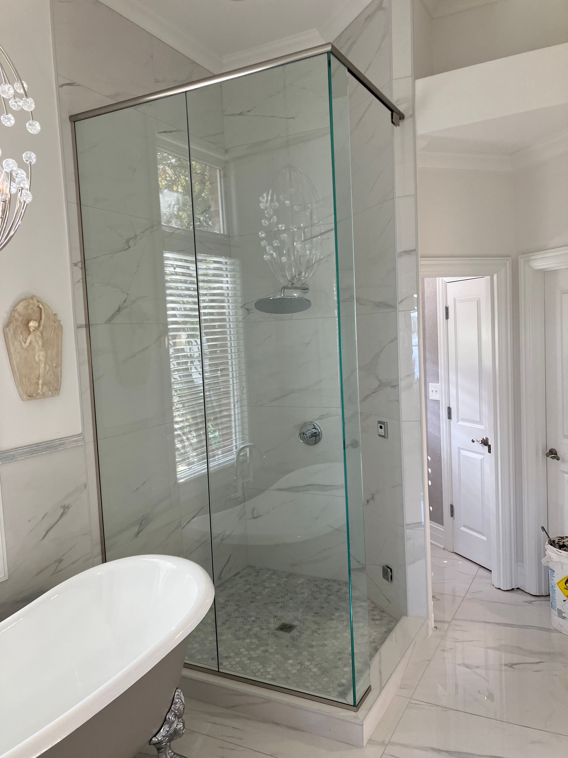 A walk-in glass shower enclosure in a marble-tiled bathroom featuring a freestanding tub and an open doorway.