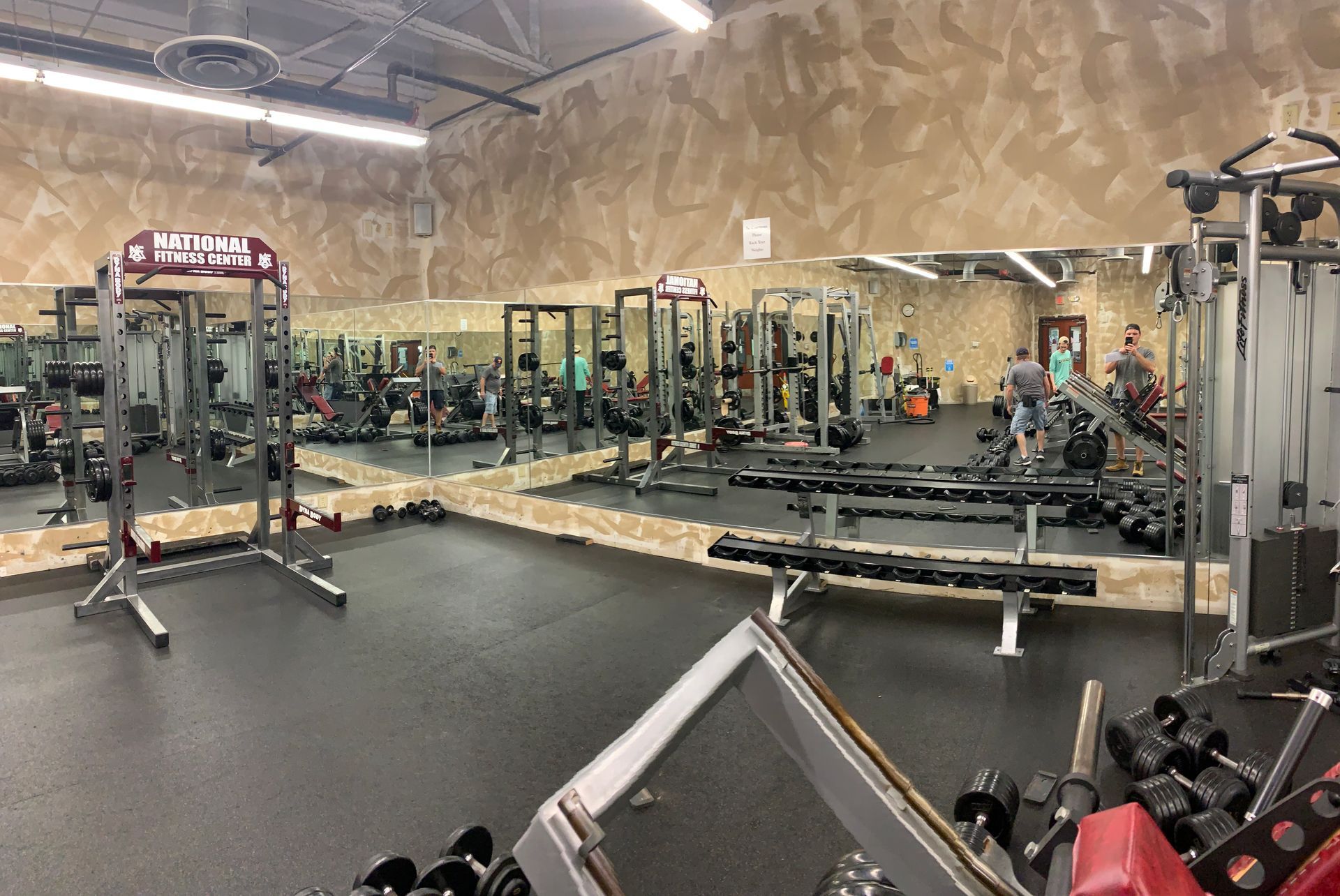 A brightly lit gym with squat racks, weight benches, and dumbbells, reflected in a large wall mirror.